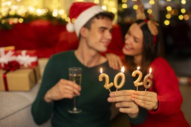 Yeni yılı kutlayan genç çiftin elindeki 2026 numaralı mumlara odaklanın ve Xmas süslü odasında ellerinde şampanya bardakları tutun.
