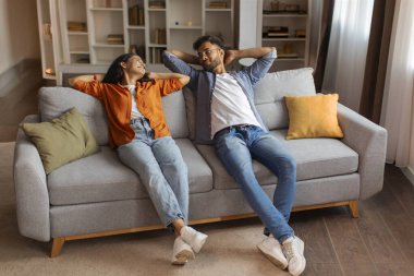 Rahat genç Hintli çift kanepede dinleniyor, başlarının arkasında el ele tutuşuyorlar, evin içinde sakin bir anın tadını çıkarıyorlar.