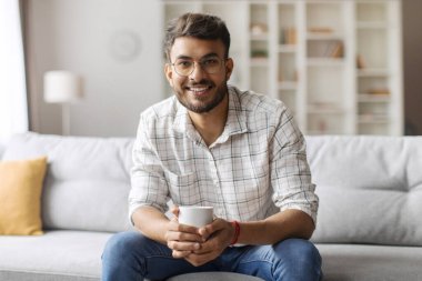Yakışıklı Hintli adam bir fincan kahveyle kanepede oturuyor ve kameraya gülümsüyor, ev eğlencesi yapıyor, mekanı kopyalıyor.