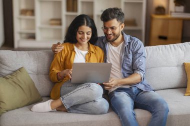 Hintli eşler kanepede dizüstü bilgisayarla rahatlarken, bilgisayarları kucaklarken ve kullanırken, internette gezinirken ya da internetten alışveriş yaparken