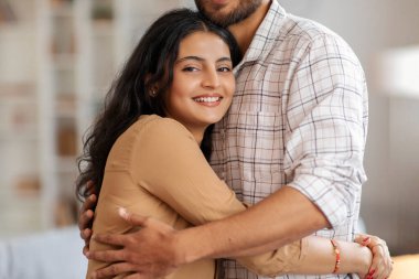 Sevecen ve mutlu Hintli çift kucaklaşıyorlar, oturma odasının içinde duruyorlar, kameraya gülümseyen kadın, boş alan. Romantik eşler sevgilerini ifade ederler.