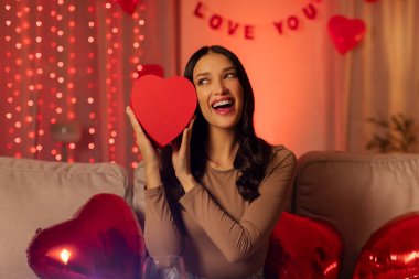 Yüzüne kalp şeklinde bir hediye tutarken gülümseyen bir kadın sevgililer gününü evde kutluyor. Kırmızı ışıklar ve kalp şeklinde balonlarla süslenmiş bir oda.