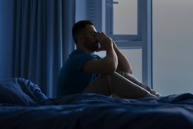 Uyku bozuklukları. Düşünceli bir adam pijamasını giymiş, geceleri yatakta oturuyor, kafaya dokunuyor, kâbuslardan uyanmış,