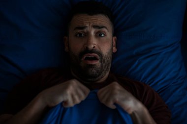 Korkmuş adam battaniyeyi tutuyor ve kameraya bakıyor, geceleri yatakta yatıyor. Erkek panik atağı, korku ve endişe. Erkek akıl sağlığı sorunları