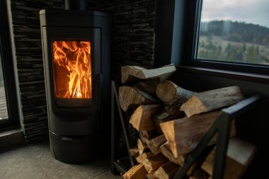 Konforlu ev odasının köşesinde, orman ve dağları gösteren geniş pencereli modern yanan sobanın yanındaki odun yığını.