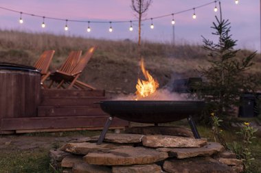 Arka bahçedeki taşların üzerinde metal ocakta yanan ateş, arka planda tahta sandalyeler ve sicim ışıkları, alacakaranlıkta yanan sahne.