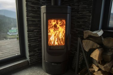 Odanın köşesinde odun sobası parlak bir şekilde yanıyor, yakınlarda kütükler yığılıyor, dağ manzaralı büyük bir pencere var.