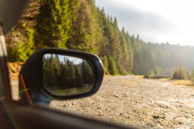 Araba yolda giderken uzun ağaçlarla çevrili, dikiz aynası yoğun ormanın yansımasını gösteriyor, güneş ışığı dağlarda parlak bir manzara yaratıyor.