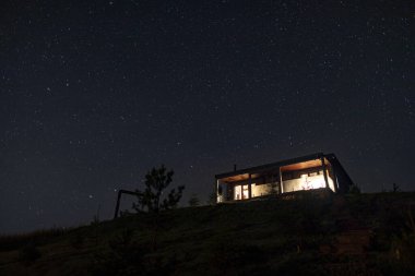 Dağlardaki ev manzarası yıldızlı gece gökyüzünün altında, pencereler aydınlık, içerideki sıcaklığı gösteriyor.
