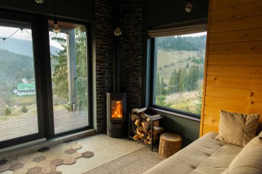 Ahşap sobalı oturma odası, tepelerin ve ağaçların manzarasını gösteren büyük panoramik pencere, kanepeli oda, ahşap duvarlar ve rahat atmosfer.