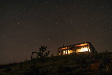 Ev, yıldızlarla dolu bir tepenin altında duruyor. Yumuşak bir ışık parlıyor. Geceleri dağlarda.