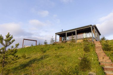 Teraslı ev kırsal ya da dağlık alanda çimenli tepenin üzerinde duruyor, gökyüzü dağınık bulutlarla, serbest alanla aydınlık.