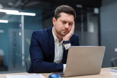 Sıkılmış ve yorgun işadamı kafasını avucunun üzerine koyup üzgün bir şekilde bilgisayar ekranına bakıyor, modern ofis içlerinde masa başında oturuyor.