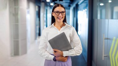 Başarılı iş kadınının portresi koridordaki modern çalışma alanında duruyor ve dizüstü bilgisayarını tutuyor, kameraya bakıyor ve gülümsüyor.