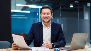 Gülümseyen iş adamı elinde belgelerle, dizüstü bilgisayarla online ticaret pazarı teknolojisi araştırması yapıyor. Ofiste evrak işleriyle uğraşan bir iş adamı.