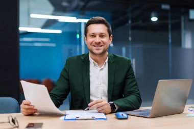 Gülümseyen iş adamı elinde belgelerle, dizüstü bilgisayarla online ticaret pazarı teknolojisi araştırması yapıyor. Ofiste evrak işleriyle uğraşan bir iş adamı.