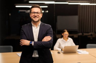 Başarılı bir iş adamı, arka planda çalışan kadın meslektaşıyla toplantı odasında duruyor. Kollarını kavuşturup kameraya gülümseyen bir adam şirketi müdürü.