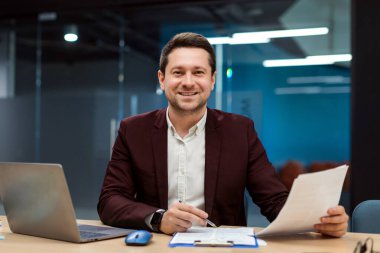 Gülümseyen iş adamı elinde belgelerle, dizüstü bilgisayarla online ticaret pazarı teknolojisi araştırması yapıyor. Ofiste evrak işleriyle uğraşan bir iş adamı.