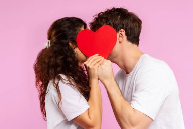 Romantik çift kırmızı kağıt kalp kartının arkasına saklanıp öpüşüyor Sevgililer Günü 'nü kutluyor pembe stüdyo arka planında poz veriyor.