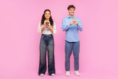 Akıllı telefonlarını kullanan, mesajlaşan, pembe stüdyo arka planında tek başına duran pozitif kadın ve erkeğin vücut uzunluğu.