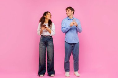 Modern internet iletişimi. Mutlu adam ve kadın cep telefonu kullanıyor, birbirlerine bakıyor ve gülümsüyorlar, pembe arka planda duruyorlar, uzun bir çekim.