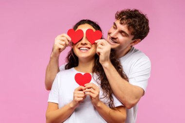 Neşeli adam, kadınların gözlerini kırmızı kağıt kalplerle kaplıyor. Pembe arka plana karşı neşeli bir an paylaşıyorlar. Sevgililer Günü