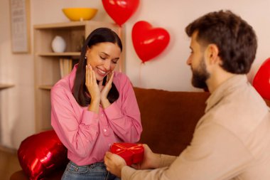 Kırmızı hediye kutusunu mutlu kadına veren adam, neşeyle gülümseyen kadın, odayı kalp balonları süslüyor. Romantik hediye, yıldönümü veya sevgililer günü kutlaması