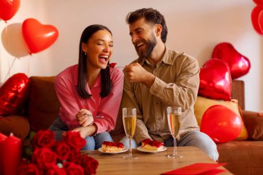 Mutlu çift tatlıyı paylaşıyor, gülümsüyor ve birbirlerini neşeyle besliyorlar, kırmızı balonlar ve güllerle süslü bir odada koltukta oturuyorlar. Yıldönümü veya Sevgililer Günü kutlaması