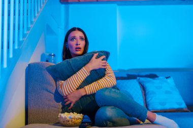 Korkmuş genç bir kadın TV 'de korku filmi seyrediyor, yastığı tutuyor ve sarılıyor, akşamları evdeki kanepede oturuyor, boşluğu kopyalıyor.