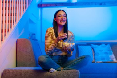 Kanepede oturup komedi filmi izleyen, abur cubur yiyen, evde film seyreden, televizyonda komik web dizileri seyreden heyecanlı bir kadın.