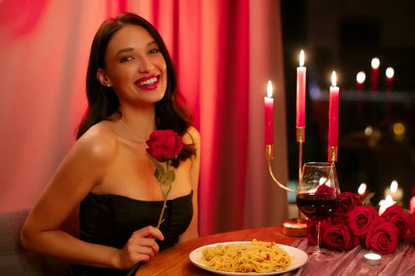 Elinde gülle kameraya gülümseyen, romantik bir akşam yemeği sırasında restoranda oturan mutlu kadın. Sevgililer Günü kutlaması