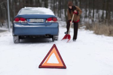 Karlı yolda arabanın yanındaki karı kaldıran kadın, ormanlık alanda kış mevsiminde uyarı üçgeniyle uyanıyor.
