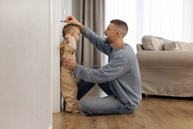 Sevgi dolu bir baba, beyaz duvarın yanında küçük bir kızın boyunu ölçüyor, çocukların kafasının üstünde elini tutuyor, oturma odasını aydınlatıyor. Büyüme İzleme Süresi