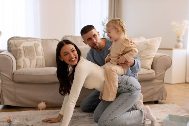 Mutlu bir aile evde eğleniyor, küçük bir kız ata biner gibi annelere biniyor, ebeveynler ve çocuklar birlikte oturma odasında vakit geçiriyorlar.