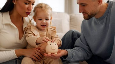 Ebeveynler küçük kızlarına kumbarayla nasıl para biriktireceğini öğretiyor, oturma odasında birlikte oturuyorlar.