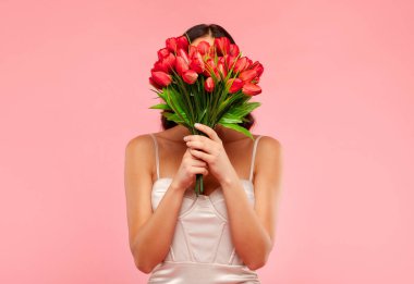Uluslararası Kadınlar Günü konsepti. Kırmızı lale buketi tutan, yüzünü kaplayan, pembe arka planda poz veren tanınmamış bir kadın.