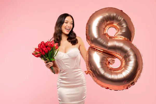 Uluslararası Kadınlar Günü. Zarif elbiseli güzel bir bayan elinde bir buket lale ve pembe arka planda sekiz numaralı balon tutuyor.