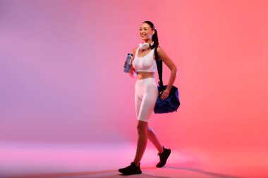 Beyaz spor giyimli ve çantalı genç bir kadın spor salonuna gidiyor. Gradyan pembe ve şeftali rengi arka planda poz veriyor. Tam boy. Egzersiz konsepti