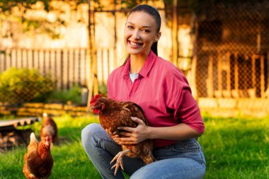 Özel çiftlikte elinde tavuk tutan, kameraya gülümseyen, çiftlikte poz veren organik kümes hayvanları yetiştiren mutlu genç bir kadının portresi.