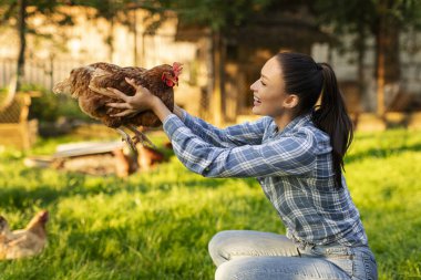 Kahverengi tavuk tutan ve gülümseyen genç bir çiftlik işçisi çiftlik arka bahçesinde poz veriyor.