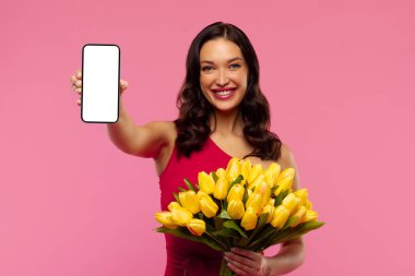 Elinde bir buket lale tutan güzel bir kadın ve boş ekranlı cep telefonunu pembe arka planda, makette, kopyalama alanında izole edilmiş olarak gösteriyor. Online teklif, mobil uygulama, dağıtım