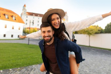İki turist eski binaların önünde mutlu bir şekilde poz veriyor ve dışarıda birlikte vakit geçiriyorlar. Adam kadını sırtında taşıyor.