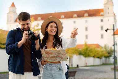 Turistler dışarıda gezmekten ve manzaralara bakmaktan hoşlanıyorlar. Kadın harita tutarken, erkek fotoğraf çekiyor.