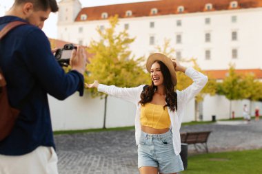 Kadın mutlu poz verirken, erkek parkta fotoğrafını çekiyor. Arka planda eski binalar, yürüyen ve şehri keşfeden bir çift.