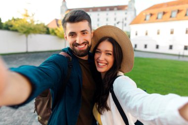 İki turist kucaklaşıp selfie çekiyor, kameraya gülümsüyor, güneşli bir günde tarihi mekanda yürüyor.