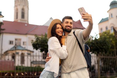 Sevgili bir çift, tarihi binanın yakınında selfie çekiyor. Erkek ve kadın telefon kameralarına sarılıp gülümsüyor.