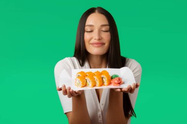 Geleneksel taze suşi rulolarını tutan ve koklayan tatmin olmuş kadın tabakta servis edilen Japon yemek aşığı iyon yeşili stüdyo arka planında poz veriyor.