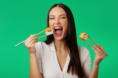 Heyecanlı bir kadın elinde suşi ruloları tutuyor ve yiyor, geleneksel Japon yemeklerini yiyor, yeşil stüdyo arka planında izole edilmiş kameraya bakıyor.