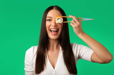 Heyecanlı kadın gözlerini çiğ suşi rulosuyla kapadı, Japon yemeklerini seviyor, yeşil stüdyo arka planında izole edilmiş poz veriyor.