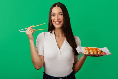 Bir elinde suşi tabağı, diğer elinde çubuklar, kameraya gülümseyen yeşil arka planda poz veren tatmin olmuş bir kadın.